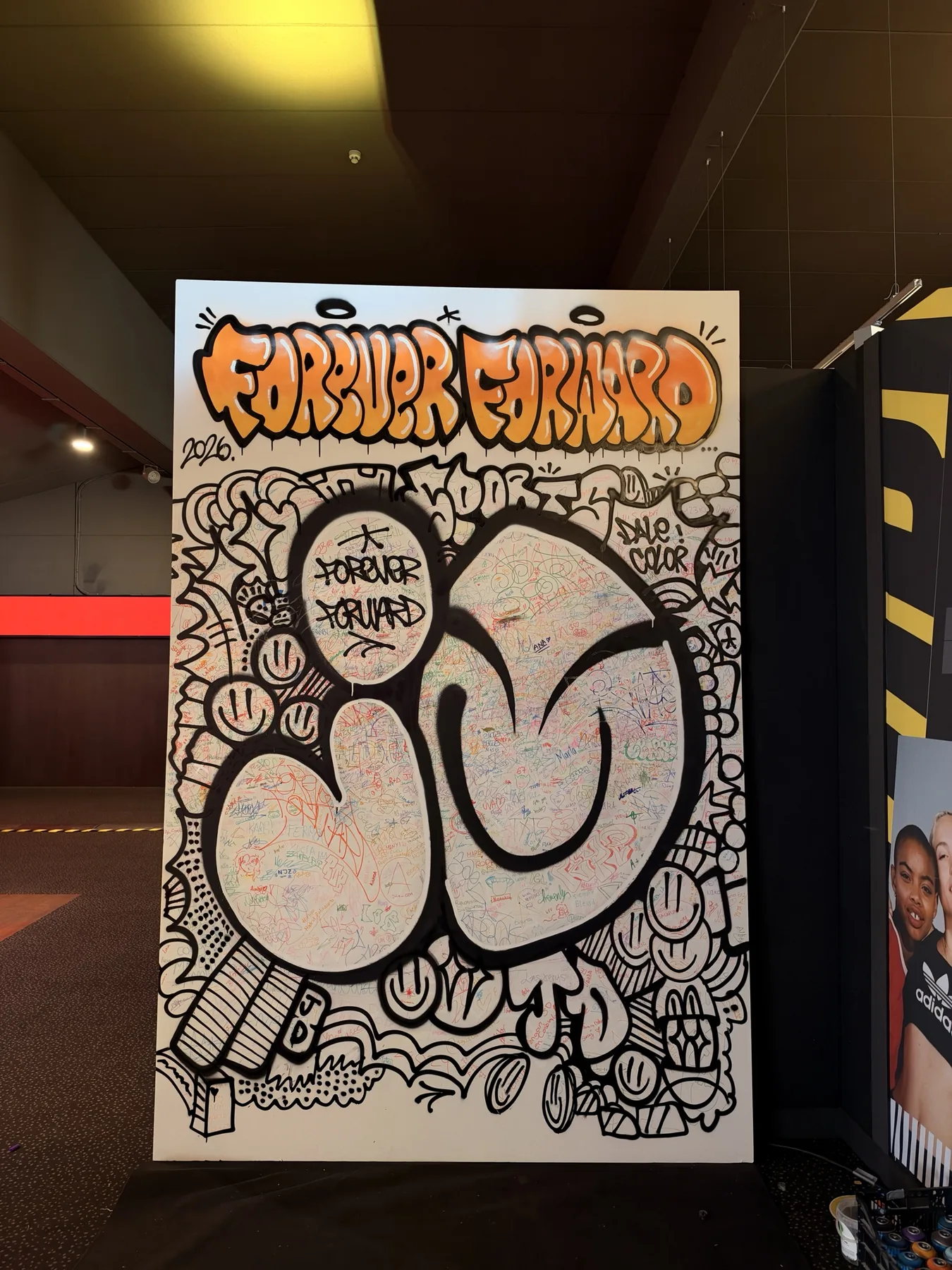 Lienzo colaborativo «Forever Forward» terminado para JD Sports: letras JD en spray negro sobre cientos de firmas de alumnos en Kinépolis Ciudad de la Imagen, Madrid.
