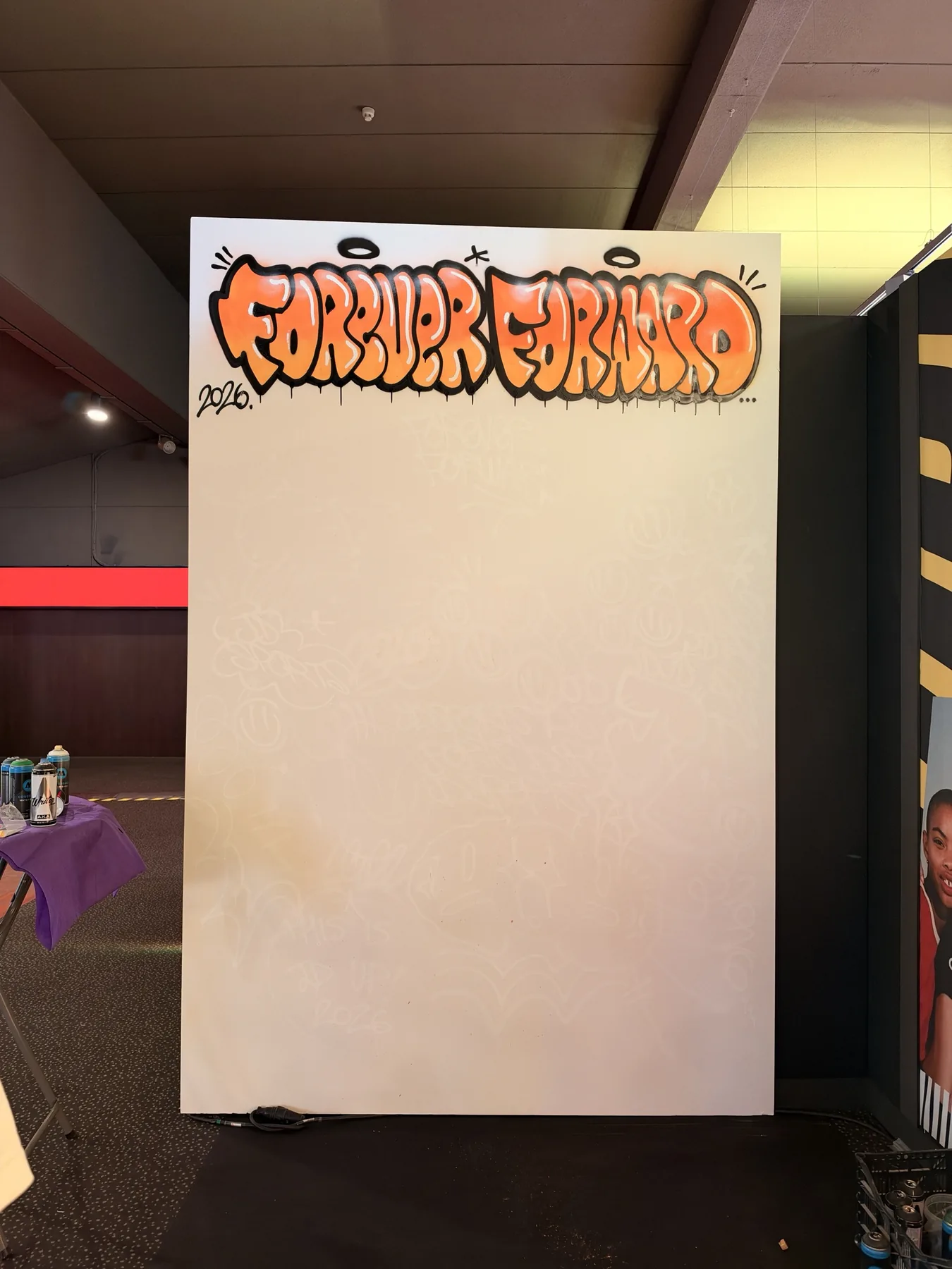 Antes: lienzo blanco con el lettering «Forever Forward 2026» pintado en spray naranja, listo para las firmas, en el stand de JD Sports en Kinépolis.