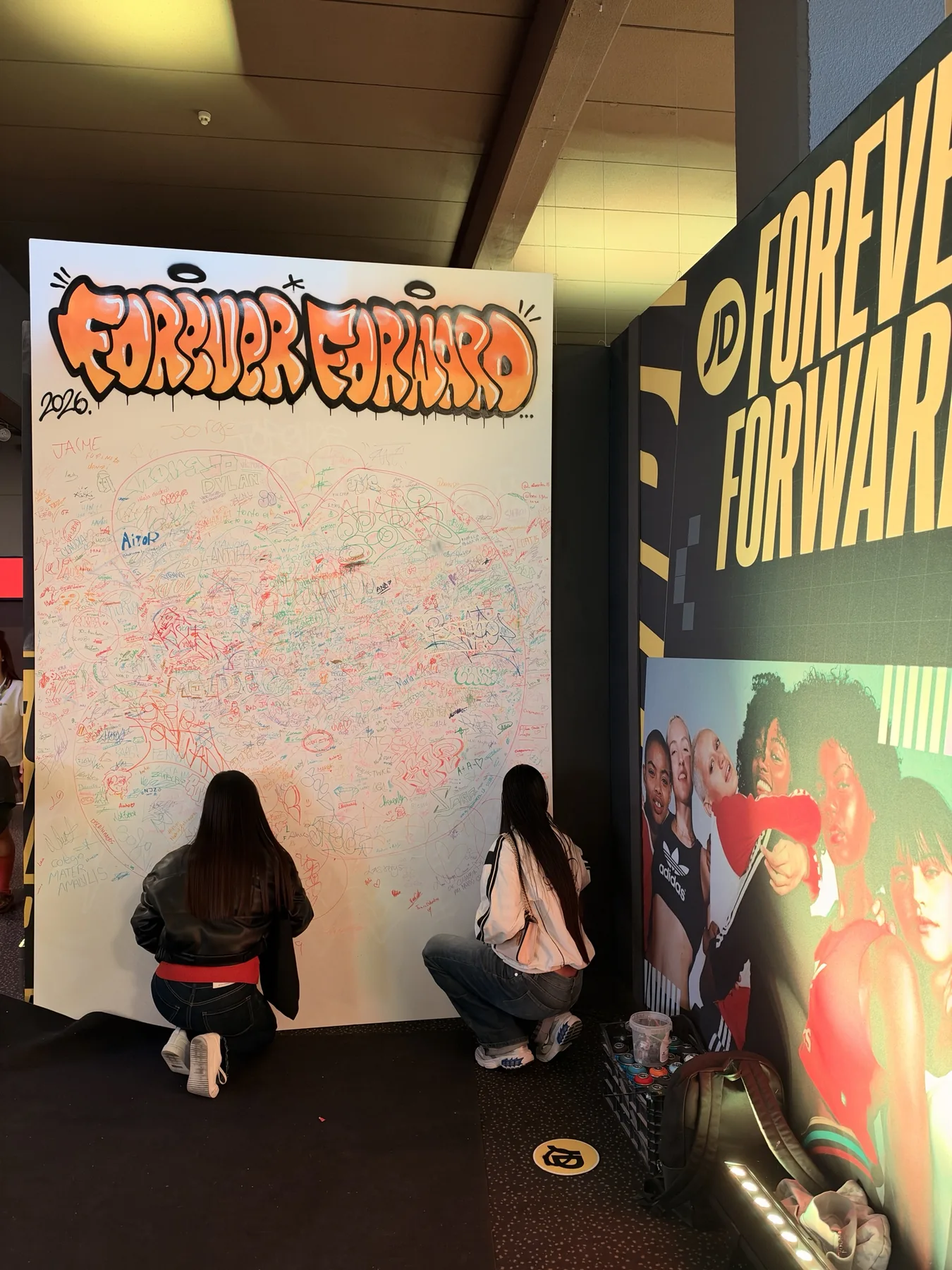 Dos alumnas firmando la parte baja del lienzo «Forever Forward», ya casi lleno, junto a la gráfica de JD Sports.