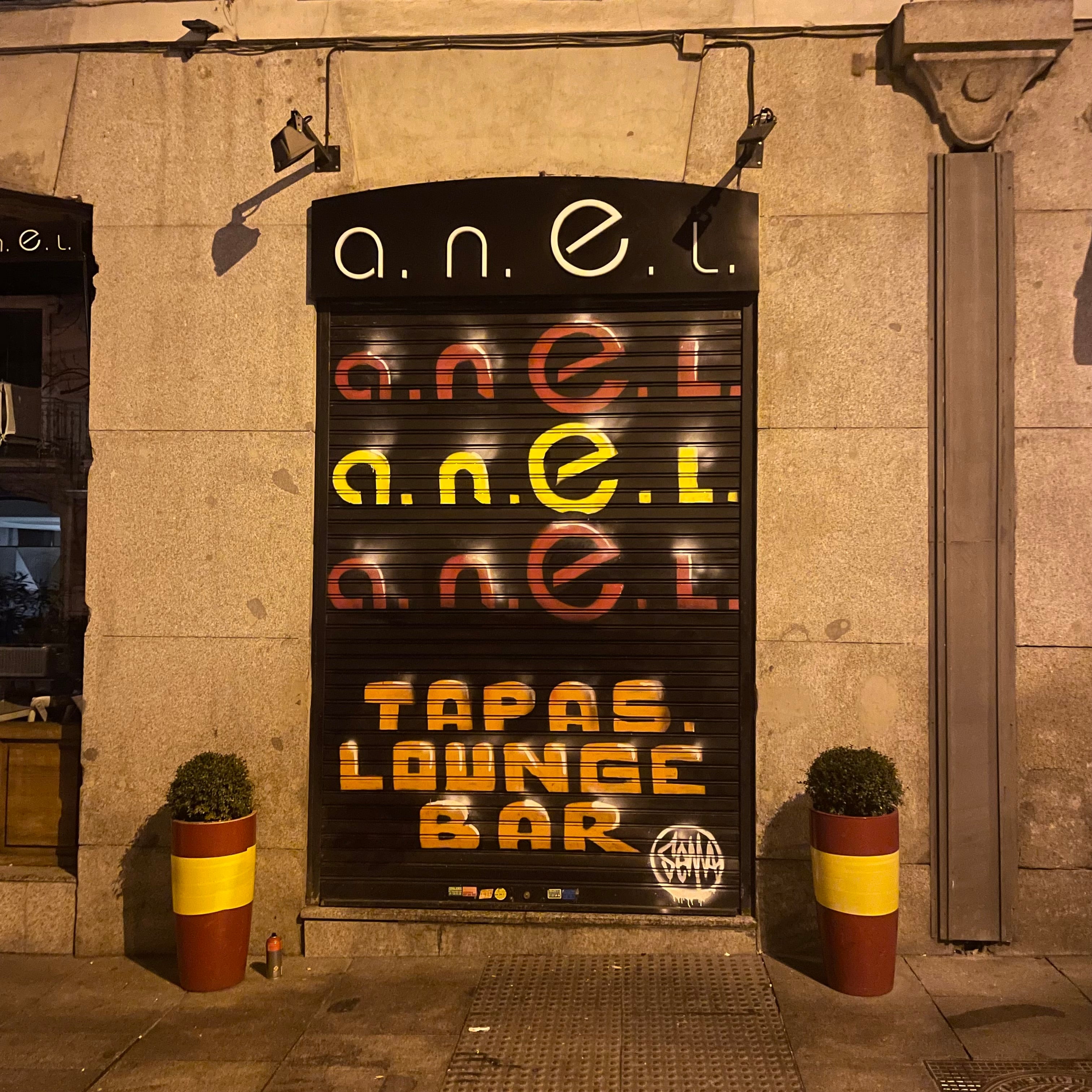 Cierre metálico de bar junto a Puerta de Alcalá pintado con logotipo A.N.E.L. formando la bandera de España y texto Tapas Lounge Bar.