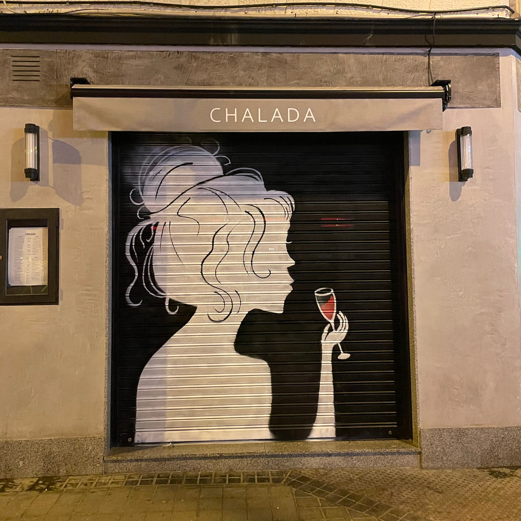 Cierre metálico de restaurante en esquina pintado con silueta de mujer y copa de vino. Diseño minimalista blanco sobre negro para bodega La Chalada.