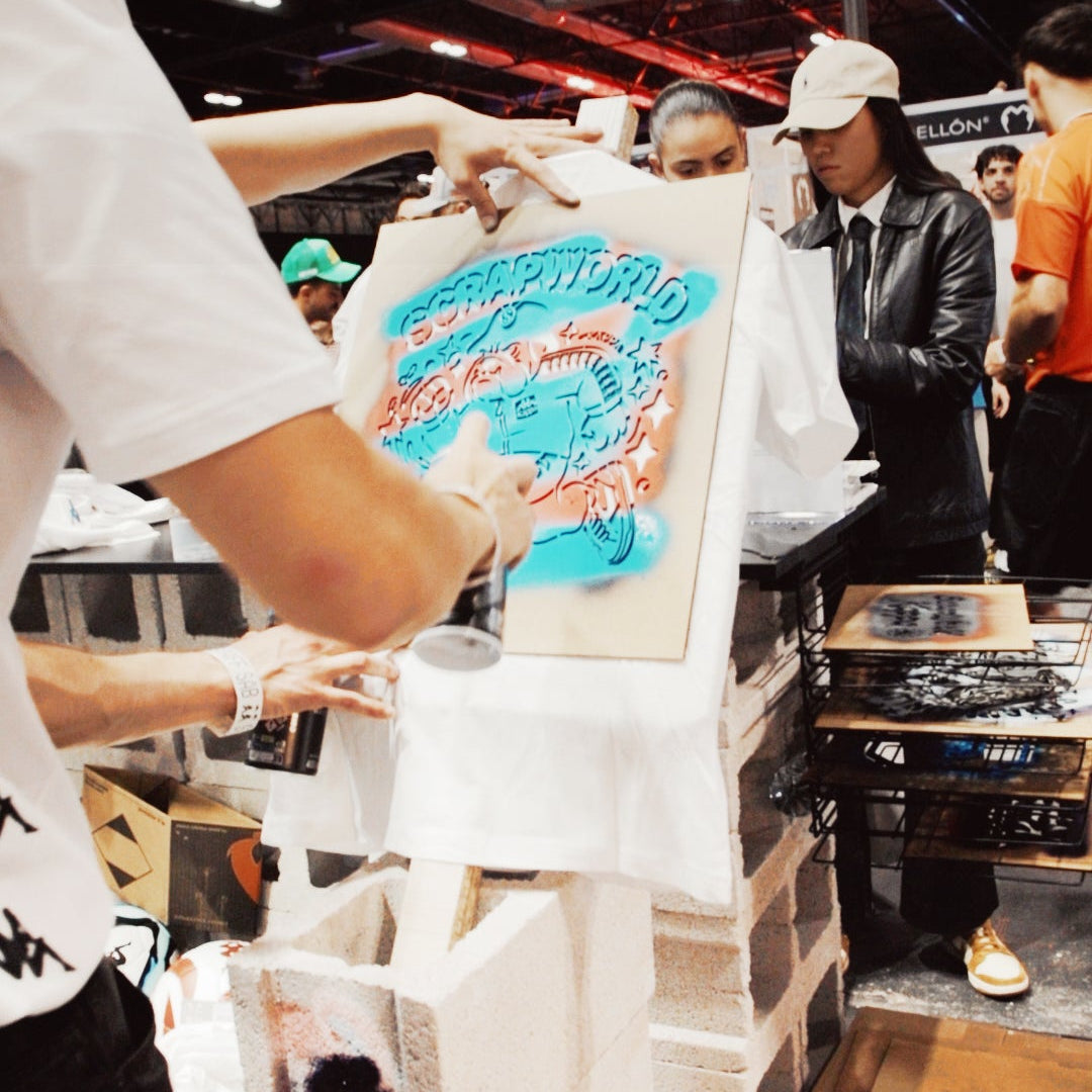 Graffitero profesional en Madrid aplicando spray base agua celeste sobre una plantilla con el logo de Scrapworld en una camiseta blanca de Kappa.