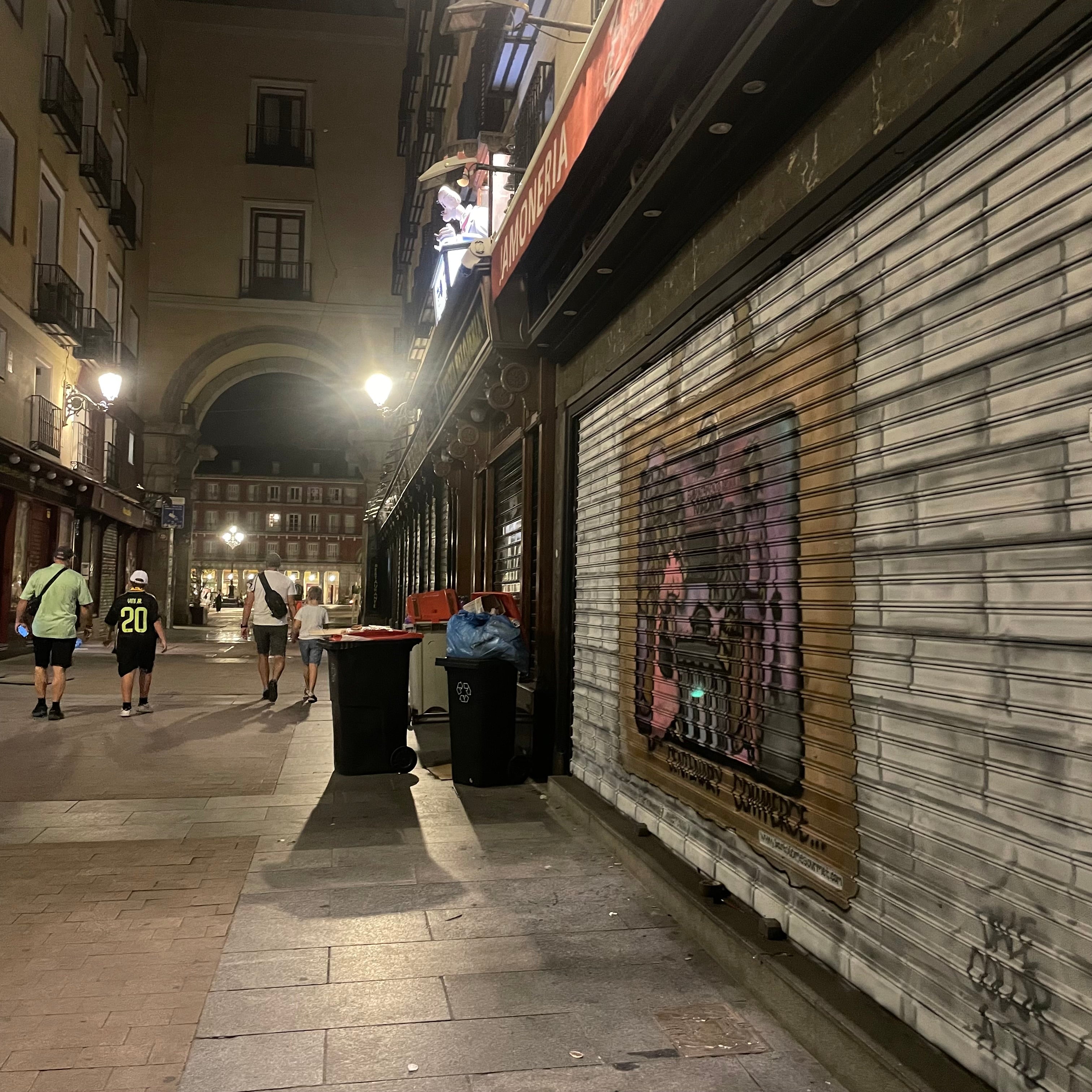Perspectiva nocturna de calle junto a Plaza Mayor. Fachada de jamonería decorada con graffiti artístico simulando ladrillo y motivos históricos.