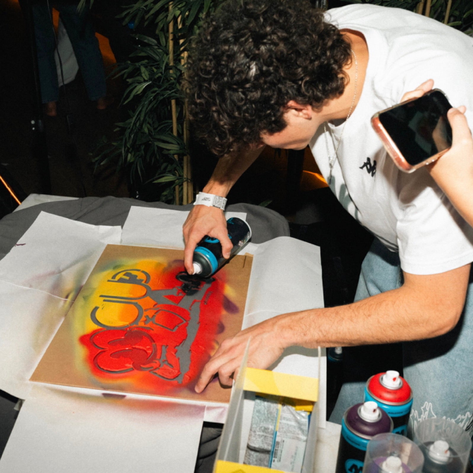 Pintores de graffiti en Madrid realizando customización en vivo. Artista aplicando spray base agua sobre plantilla para diseño de camisetas de Fraeternity en evento de Panda Club.